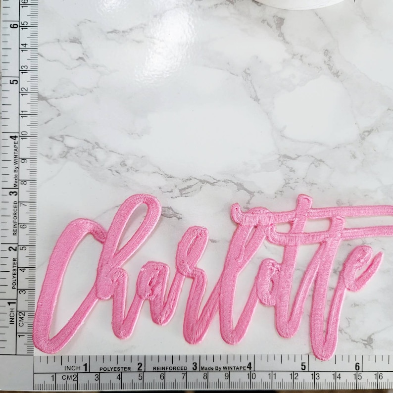 May include: A pink embroidered patch featuring the name Charlotte in a cursive script font. The patch is positioned next to a measuring tape showing a width of approximately 6 inches and a height of approximately 2.5 inches.