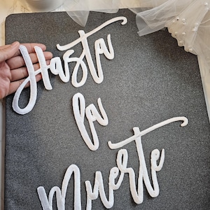 May include: White script lettering cutouts spelling the phrase Hasta la Muerte. The letters are constructed from a white material with a stitched border, arranged in three lines on a dark gray textured surface.