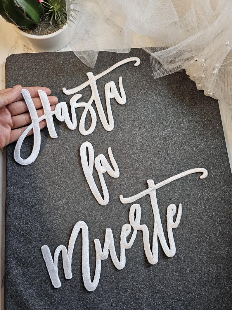 May include: White script lettering cutouts spelling the phrase Hasta la Muerte. The letters are constructed from a white material with a stitched border, arranged in three lines on a dark gray textured surface.