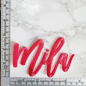 May include: A bright red embroidered patch featuring the name Mila in a cursive script font. The patch is positioned next to a measuring tape showing a width of approximately 5 inches and a height of approximately 2.5 inches.