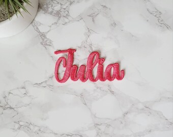 May include: Pink embroidered iron-on patch with the name "Julia" in a cursive font.
