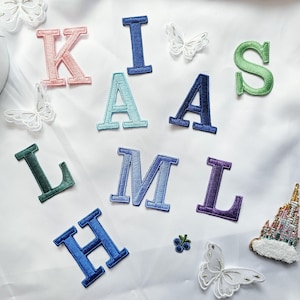 May include: A collection of embroidered fabric patches featuring various letters of the alphabet in colors including pink, light blue, dark blue, green, and purple, alongside decorative white butterfly patches and a small castle patch.