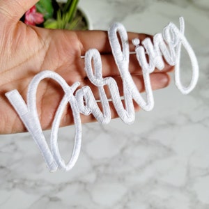 May include: A white embroidered patch featuring the word darling in a cursive script font. The lettering is stitched with a dense white thread, creating a raised texture and a clean, bright appearance against the background.