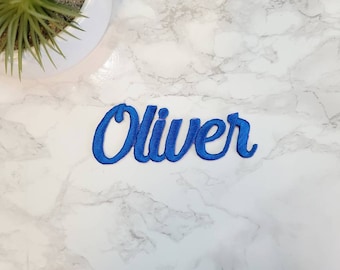 May include: Blue embroidered iron-on patch with the name "Oliver" in a cursive font.