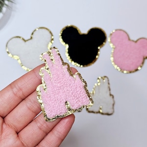 Puede incluir: Cinco parches de fieltro rosa y blanco con ribete de purpurina dorada. Los parches tienen forma de orejas de Mickey Mouse y un castillo.