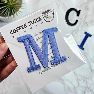 Puede incluir: Un parche de la letra M bordado en azul. El parche está sobre un fondo blanco con el texto "Coffee Juice Studio" y "Custom Apparel, Embroidery, Patches, Digital Cut, Graphic Design, & Stitch".