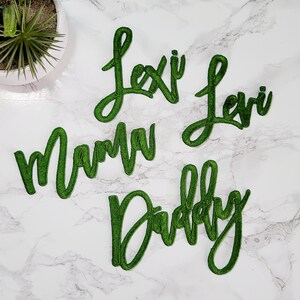 May include: Four green embroidered patches featuring cursive text. The patches display the words Lexi, Levi, Mama, and Daddy in a script font with a raised, textured stitch finish. Each patch is a separate piece with a distinct green thread border.