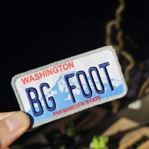Puede incluir: Un parche rectangular que se asemeja a una placa de matrícula del estado de Washington. El parche presenta las palabras "BG FOOT" en azul, con "WASHINGTON" en rojo arriba y "EVERGREEN STATE" en rojo abajo. El parche tiene un fondo blanco con un gráfico de montaña azul.