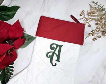 May include: A white quilted Christmas stocking featuring a deep red cuff and a dark green embroidered letter A in the centre. The stocking includes a matching red fabric loop at the top corner for hanging purposes.