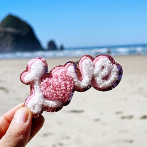 May include: A pink and white felt patch with the word "LOVE" in a fluffy, cursive font. The patch is held in a hand against a blurred background of a beach and ocean.