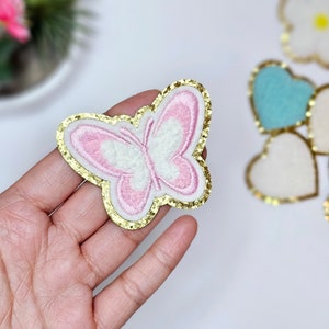 May include: A pink and white butterfly applique with a gold glitter outline. The butterfly is on a hand.