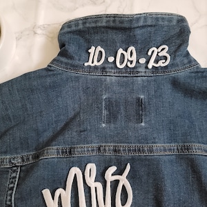 May include: A blue denim jacket with white embroidery. The back of the jacket says "Mrs" and the collar has the date "10.09.23" embroidered on it.