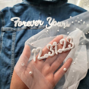 Op de afbeelding: Een witte tule stof met parelversieringen en de datum "12.31.22" geborduurd in witte draad. De stof wordt in een hand gehouden.