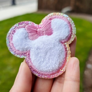 Può includere: Applique in feltro rosa e bianco a forma di testa di topo dei cartoni animati con un fiocco rosa. L'applique ha un bordo scintillante iridescente.