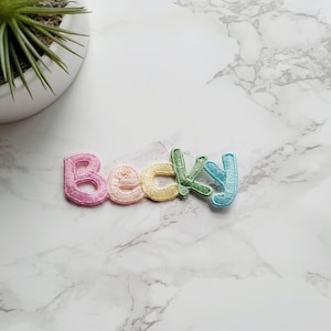 May include: The name "Becky" spelled out in colorful embroidered letters on a white marble background.