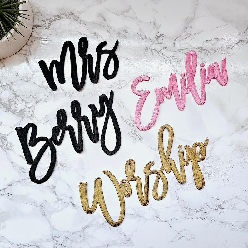 May include: Three embroidered fabric patches in cursive script. One black patch reads Mrs, another black patch reads Berry, one pink patch reads Emilia, and one gold patch reads Worship. The lettering features a raised, stitched texture.