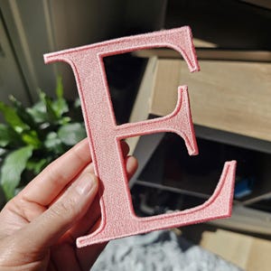 May include: A light pink embroidered letter E patch featuring a clean, block-style font with a stitched border. The fabric surface has a textured, thread-based finish, and the edges are clearly defined by the surrounding embroidery stitching.