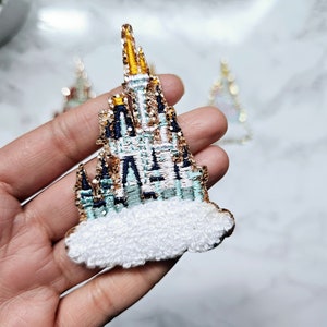 Puede incluir: Una nube blanca y esponjosa con un castillo bordado en oro, azul y verde en la parte superior. El castillo tiene una alta aguja puntiaguda y múltiples torres. El castillo es un símbolo popular de un famoso parque de atracciones.