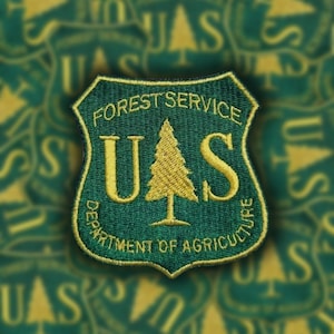 May include: A green and gold embroidered patch with the text "FOREST SERVICE US DEPARTMENT OF AGRICULTURE" and a stylized pine tree.