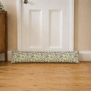 May include: A long, rectangular fabric door draught excluder featuring a repeating botanical pattern of green leaves and thin brown stems on a cream-coloured background, positioned horizontally along the base of a white panelled door.