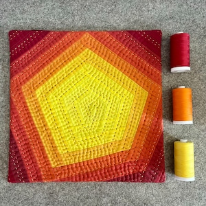 May include: A quilted cushion with a geometric design in shades of red, orange, and yellow. The cushion is surrounded by spools of thread in matching colours.