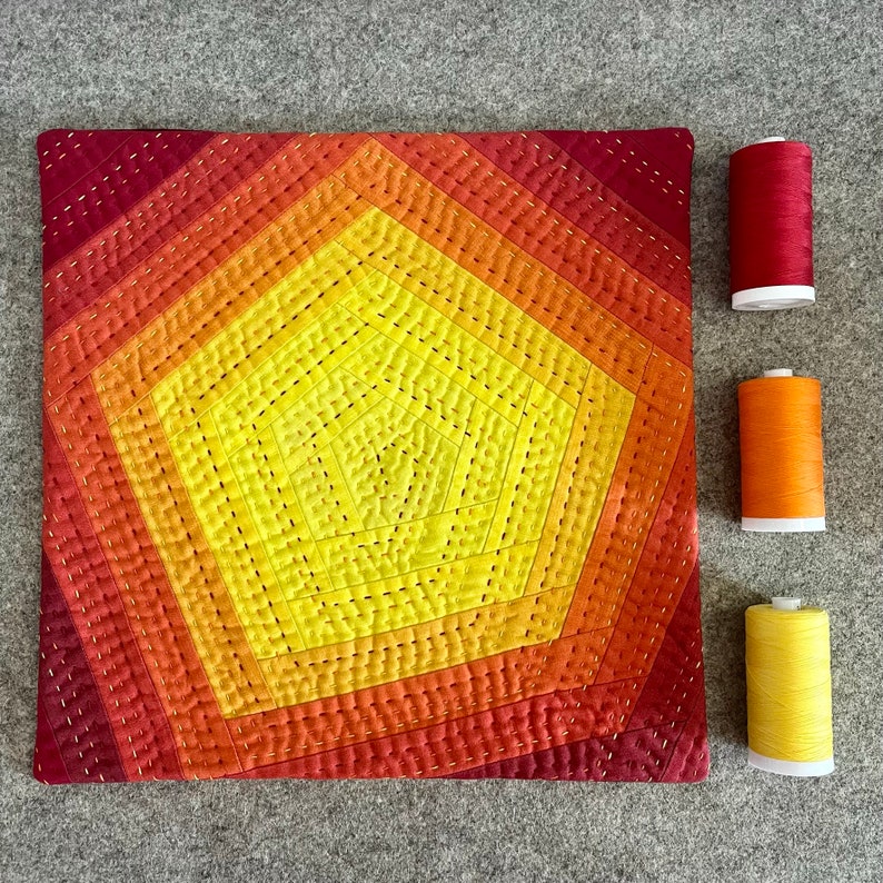 May include: A quilted cushion with a geometric design in shades of red, orange, and yellow. The cushion is surrounded by spools of thread in matching colours.