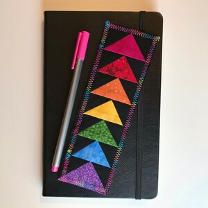 May include: A black leather journal with a rainbow fabric bookmark. The bookmark is made of sewn triangles in a rainbow pattern.