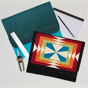 May include: A teal fabric laptop sleeve with a ruler and a pair of scissors. A black and white notebook with a grid pattern is visible in the background. A black and red patchwork quilt with a geometric design is on top of the sleeve.