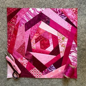 May include: A pink and purple patchwork quilt with a spiral design. The quilt is made of various fabrics with different patterns, including floral, geometric, and marbled. The quilt is edged with a white border.