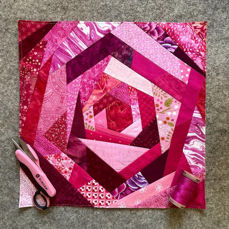 May include: A pink and purple patchwork quilt with a spiral design. The quilt is made of various fabrics with different patterns, including floral, geometric, and marbled. The quilt is edged with a white border.