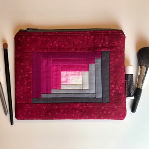 May include: A red quilted makeup bag with a log cabin pattern in shades of pink, gray, and white. The bag has a zipper closure.