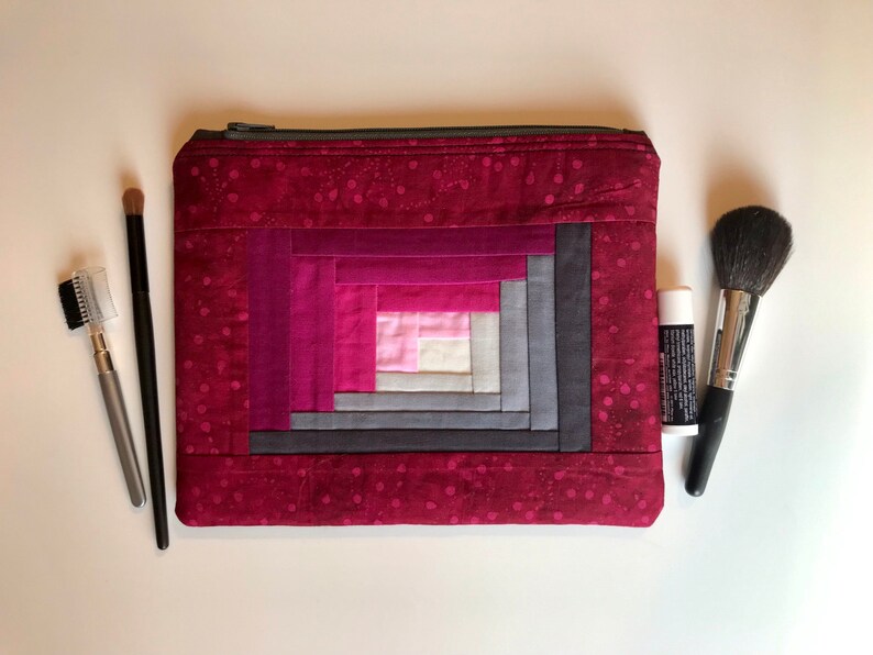 May include: A red quilted makeup bag with a log cabin pattern in shades of pink, gray, and white. The bag has a zipper closure.