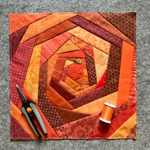 May include: A quilt with a spiral pattern in shades of orange, brown, and red. The quilt is made of fabric scraps sewn together in a patchwork design. A pair of scissors and a spool of thread are lying on top of the quilt.