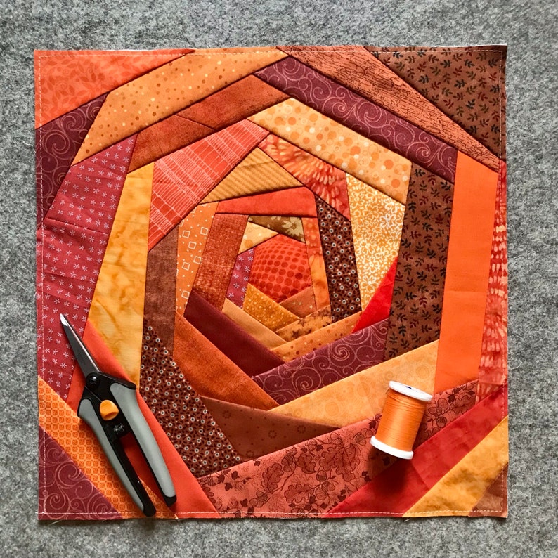 May include: A quilt with a spiral pattern in shades of orange, brown, and red. The quilt is made of fabric scraps sewn together in a patchwork design. A pair of scissors and a spool of thread are lying on top of the quilt.