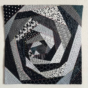 May include: A black and white quilt with a spiral pattern. The quilt is made of different fabrics with various textures and patterns. The quilt is stitched together with black thread.