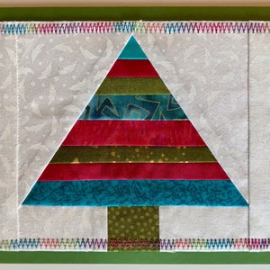 May include: A colorful patchwork Christmas tree made of fabric scraps. The tree is layered with red, green, blue, and gold fabric. The tree is surrounded by a white background with a subtle pattern.