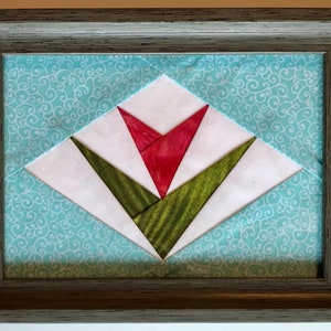 May include: A framed quilt block with a red and green tulip design. The quilt block is set against a blue background with a white swirl pattern.