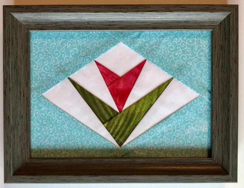 May include: A framed quilt block with a red and green tulip design. The quilt block is set against a blue background with a white swirl pattern.