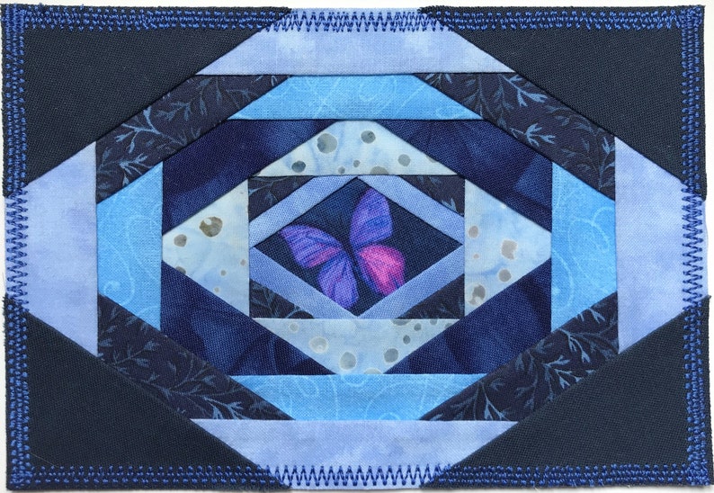 May include: A blue and purple quilt block with a butterfly design in the center. The quilt block is made of various shades of blue fabric and has a dark blue border.