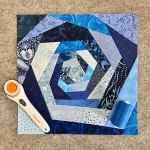 May include: A blue and white quilt block featuring a spiral design. The block is constructed from various fabric pieces, all stitched together to form a swirling pattern. The block is edged with a white border.