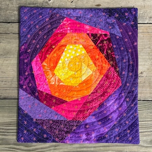 May include: A purple, pink and orange quilt with a spiral design. The quilt is made of fabric scraps and is quilted with a star-shaped stitch.