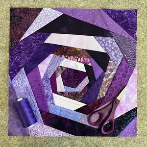 May include: A purple and white quilt with a spiral design. The quilt is made of various fabrics with different patterns. A spool of purple thread and a pair of purple scissors are on the quilt.