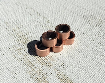 May include: A stack of six copper rings, each with a small gap. The rings have a shiny, metallic finish and are arranged on a textured, off-white surface. The rings are likely components for jewelry or other craft projects.