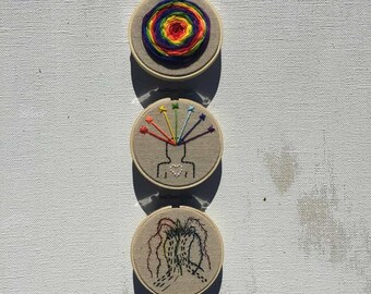 May include: Three embroidered hoops with different designs. The top hoop has a rainbow swirl design. The middle hoop has a rainbow radiating from a head with a heart in the chest. The bottom hoop has a black and white design of a tree.