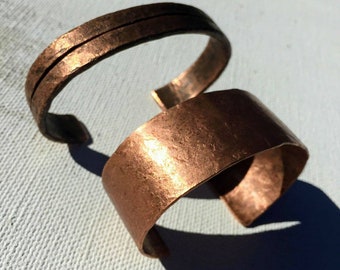 May include: Two handcrafted copper bracelets. One is a narrow band with two parallel lines etched into the surface. The other is a wide cuff with a textured finish. Both have a warm, reddish-brown color and a hammered appearance.