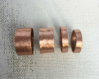 May include: Four copper rings of varying widths are displayed. The rings have a hammered texture and a warm, reddish-brown color. The rings are arranged in a row, showcasing their different sizes and designs.