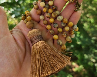 May include: A brown and yellow gemstone mala necklace with a tassel. The necklace has a gold Om charm.
