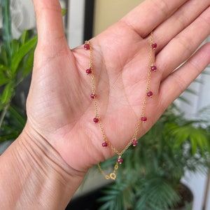 May include: A gold chain necklace with small red beads hanging from it. The necklace is on a person's hand.
