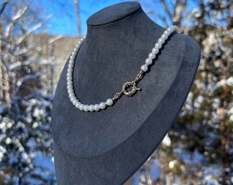 May include: A white pearl necklace with a gold clasp on a gray velvet jewelry display stand. The stand is made of wood and has a round base.