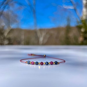 Pode incluir: Uma pulseira delicada com uma série de contas de pedras preciosas azuis e rosas, intercaladas com pequenas contas douradas. A pulseira é feita com um cordão fino, de cor bordô, com fecho de nó deslizante ajustável.